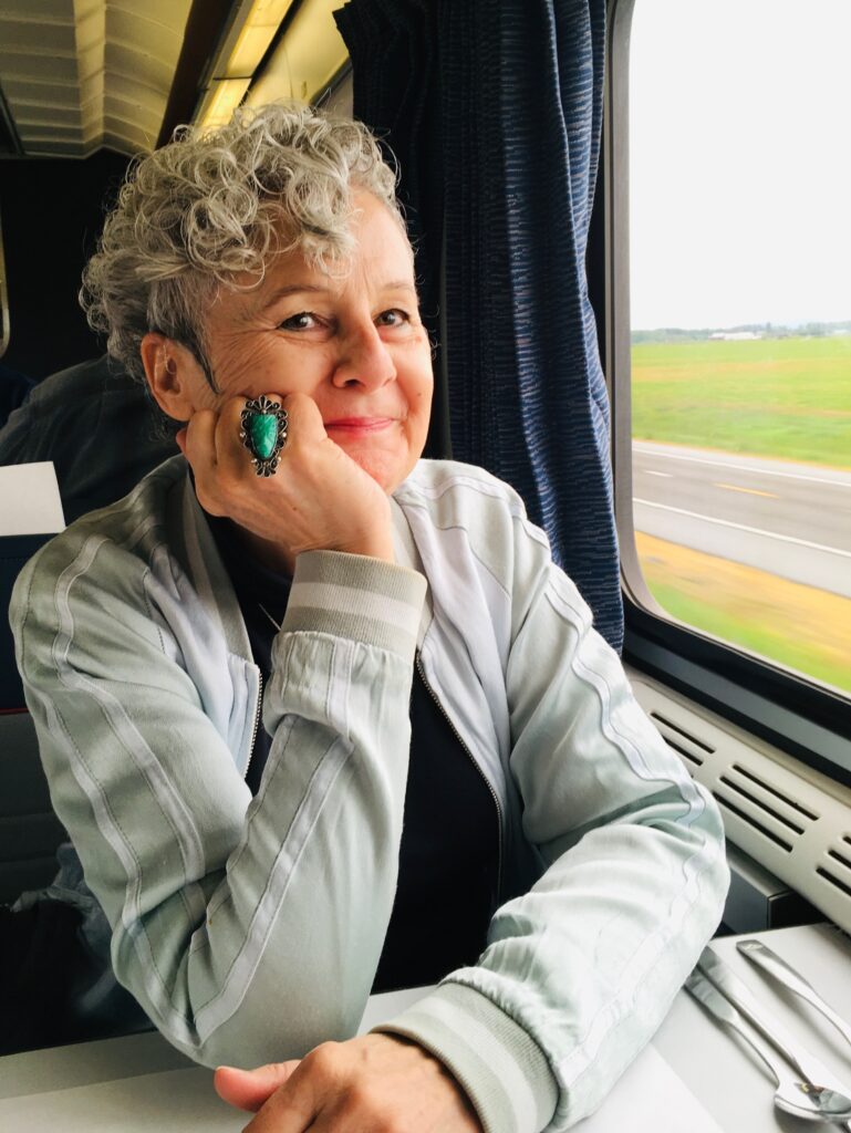 Photo of Elizabeth Wheeler on an Amtrak train next to a window, smiling at the camera, her face propped in her right hand. She has twinkly eyes and curly silver hair in a queer haircut. She is wearing a big green jade ring, hearing aids, and a celadon baseball jacket. There is a spoon on the table, which is good, because she always needs extra spoons! 