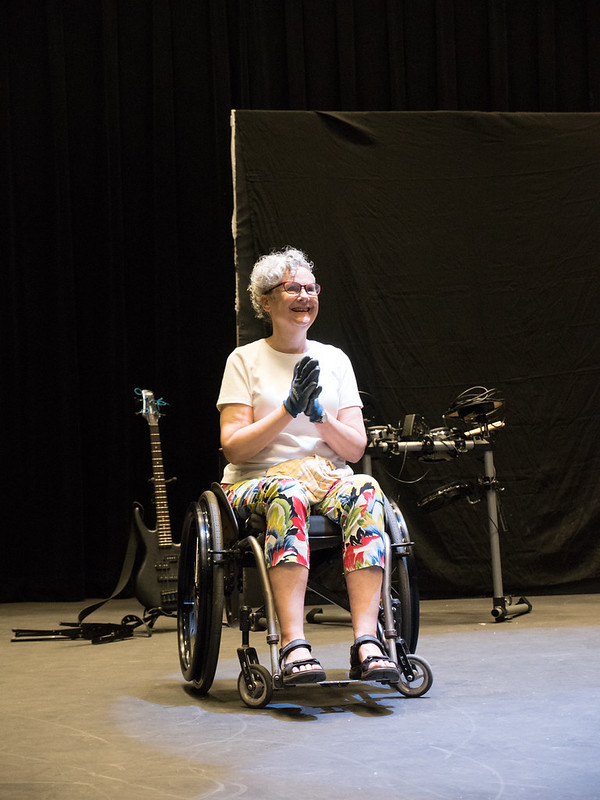 Photo of Elizabeth Wheeler onstage, rallying cast members ahead of a dress rehearsal. Elizabeth has white hair. She is wearing glasses, a white t-shirt, colorful capri pants, and wheelchair gloves, and is sitting on a manual wheelchair. She is smiling broadly at the troupe, fomenting and responding to their enthusiasm.